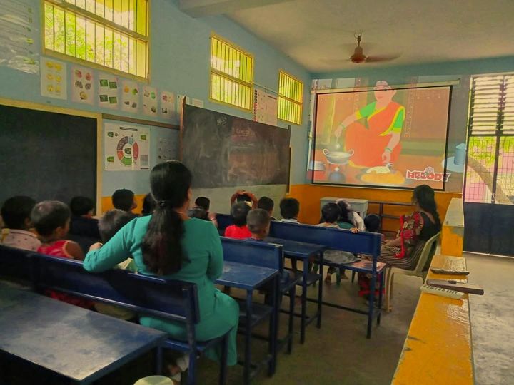 Children and teachers on classroom benches watching an animated video on a large projector screen